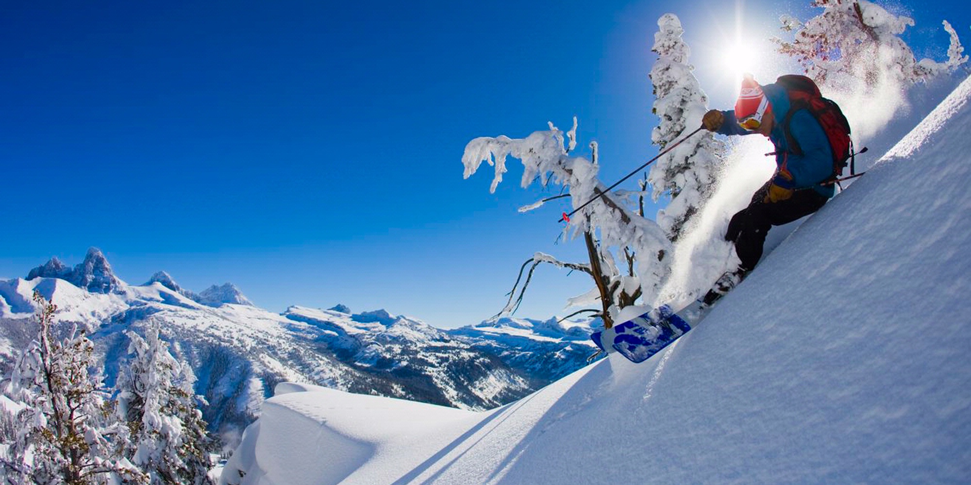 Skier carving through fresh powder on a steep slope with the snow-covered Grand Teton peaks in the background under a clear blue sky near Jackson Hole, Wyoming.